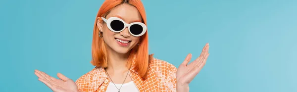 Positive, junge asiatische Frau mit gefärbten Haaren in lässiger Kleidung und Sonnenbrille, gestikulierend mit den Händen auf leuchtend blauem Hintergrund, orangefarbenem Hemd, Halskette, Generation z, rotem Haar, Banner — Stockfoto