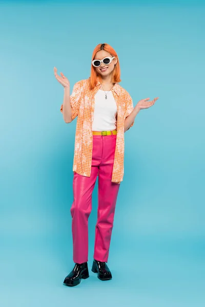 Estilo pessoal, mulher asiática feliz com cabelo tingido em pé em traje casual e óculos de sol, gesticulando com as mãos em fundo azul vibrante, camisa laranja, geração z, cabelo vermelho — Fotografia de Stock