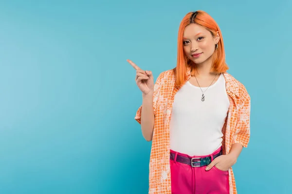 Demonstrating, young asian woman with dyed red hair standing with hand in pocket of pink pants on vibrant blue background, orange shirt, pointing with finger, showing something, generation z — Stock Photo