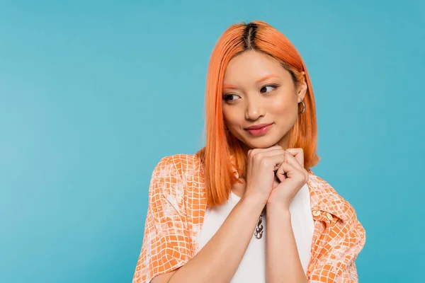 Adorable, cheerful young asian woman with dyed red hair smiling and holding hands near face on vibrant blue background, pleased, generation z, casual attire, looking away, young culture — Stock Photo