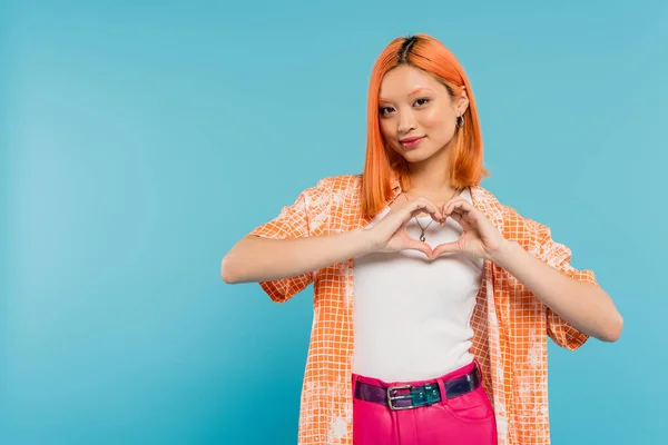 Love sign, happy face, cheerful young asian woman with dyed red hair smiling and showing heart with hands on vibrant blue background, generation z, casual attire, looking at camera, young culture — Stock Photo