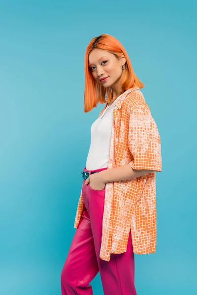 Hand in pocket, pretty and young asian woman with dyed hair standing in orange shirt and posing on blue background, looking at camera, pink pants, generation z, modern style — Stock Photo