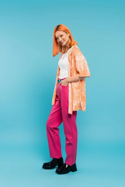 Radiant smile, full length, pretty and young asian woman with dyed hair  posing with hand in pocket on blue background, looking at camera, pink pants, generation z, modern style — Stock Photo