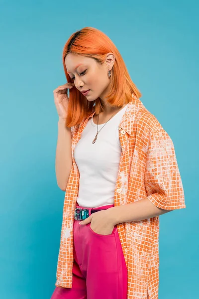 Mano en el bolsillo, sensual y joven mujer asiática con el pelo teñido de pie en camisa naranja y posando sobre fondo azul, mirando hacia otro lado, pantalones de color rosa, generación z, estilo moderno - foto de stock