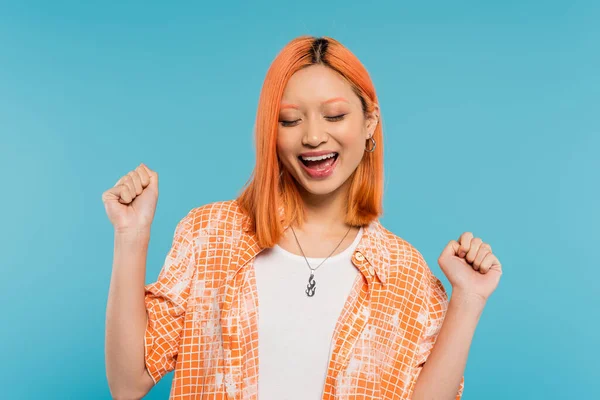 Cheering and gesturing, happy and young asian woman with dyed hair standing in orange shirt with opened mouth on blue background, closed eyes, positivity, generation z, modern style — Stock Photo