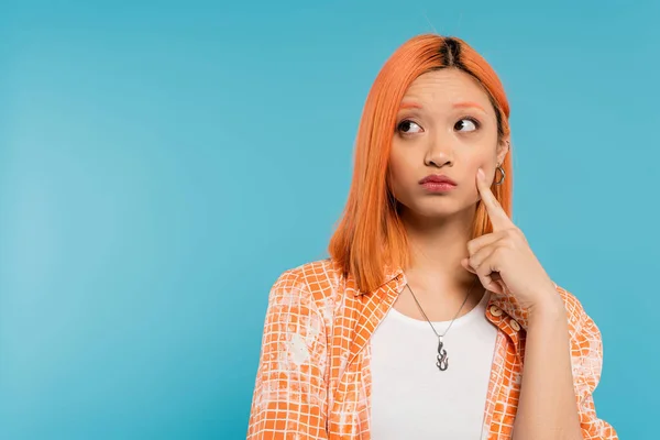 Mujer asiática pensando y mirando hacia otro lado, modelo de moda joven tocando mejilla con el dedo sobre fondo azul, pensativo, camisa naranja, generación z, colores vibrantes, cara dudosa - foto de stock