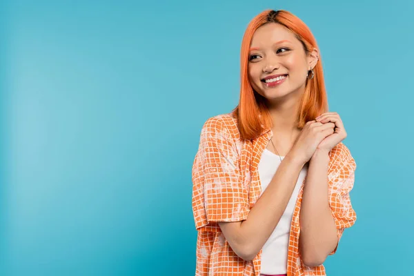 Felicidade, jovem mulher asiática com cabelo tingido em pé na camisa laranja e posando com as mãos apertadas no fundo azul, olhando para longe, alegre, adorável, geração z, estilo moderno — Fotografia de Stock