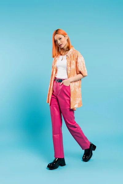 Hand in pocket, attractive and young asian woman with dyed hair standing in orange shirt and posing on blue background, looking at camera, pink pants, generation z, modern style, full length — Stock Photo