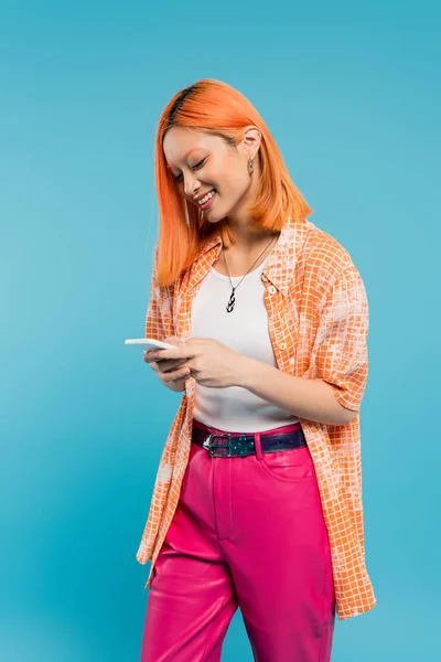 Соціальні мережі, весела азіатська жінка з пофарбованими повідомленнями про волосся, використовуючи смартфон, стоячи на синьому фоні, усміхнена, помаранчева сорочка, повсякденний одяг, цифровий рідний, покоління z — стокове фото