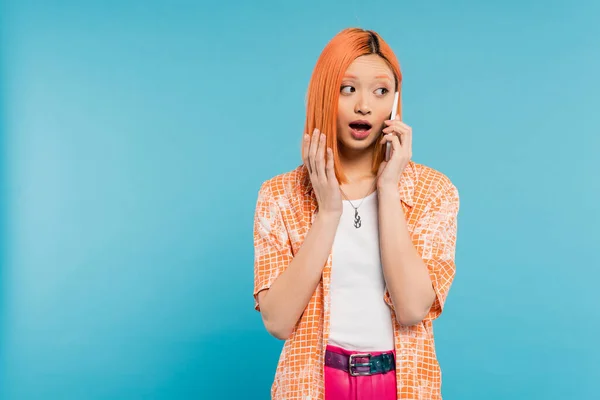 Überraschte asiatische Frau beim Telefonat, junges Model mit gefärbten Haaren steht mit geöffnetem Mund und spricht auf Smartphone auf blauem Hintergrund, schaut weg, emotionales, schockiertes Gesicht — Stockfoto