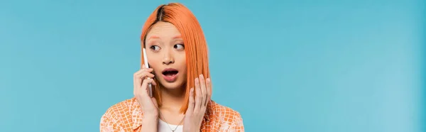 Шоковане обличчя, здивована азіатська жінка під час телефонного дзвінка, молода модель з фарбованим волоссям, що стоїть з відкритим ротом і розмовляє на смартфоні на синьому фоні, дивлячись далеко, емоційний, банер — стокове фото