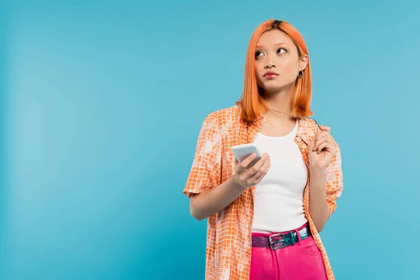 Social media influencer, pensive asian woman with dyed hair holding smartphone and looking away on blue background, mobile phone, youth culture, digital age, generation z — Stock Photo