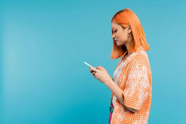 Социальный медиа влияния, молодая азиатская женщина с окрашенными волосами с помощью смартфона на синем фоне, мобильный телефон, молодежная культура, цифровой век, поколение z, обмен сообщениями, вид сбоку — стоковое фото