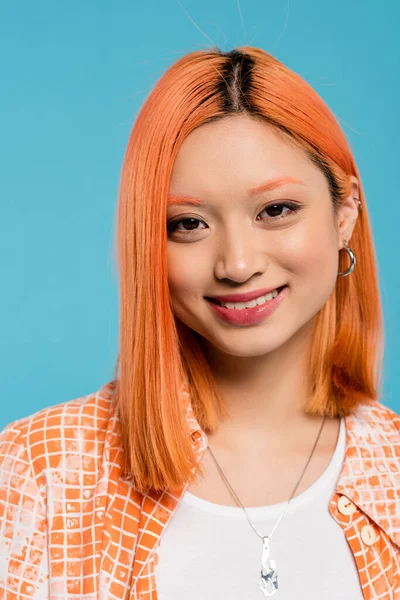 Retrato, mujer asiática joven con el pelo corto y teñido, maquillaje natural y pendientes de aro mirando a la cámara sobre fondo azul, camisa naranja, generación z, moda, cara feliz, sonrisa radiante - foto de stock