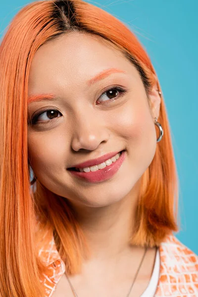 Портрет, радостная азиатская женщина с короткими и окрашенными волосами, натуральный макияж и обруч серьги глядя в сторону на голубом фоне, оранжевая рубашка, поколение z, мода, счастливое лицо, сияющая улыбка — стоковое фото