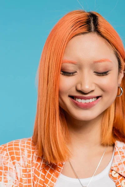 Retrato, cara alegre, joven mujer asiática con pelo corto y teñido, maquillaje natural y pendientes de aro sonriendo sobre fondo azul, camisa naranja, generación z, felicidad, emoción de alegría - foto de stock