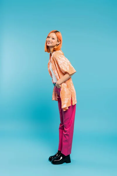 Posing with hand in pocket, joyful and young asian woman with dyed hair standing in casual wear on blue background, looking at camera, pink pants, generation z, modern style, full length — Stock Photo
