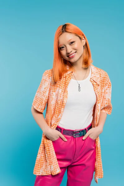 Positivity, posing with hands in pockets, joyful and young asian woman with dyed hair standing in casual wear on blue background, looking at camera, pink pants, generation z, modern style — Stock Photo
