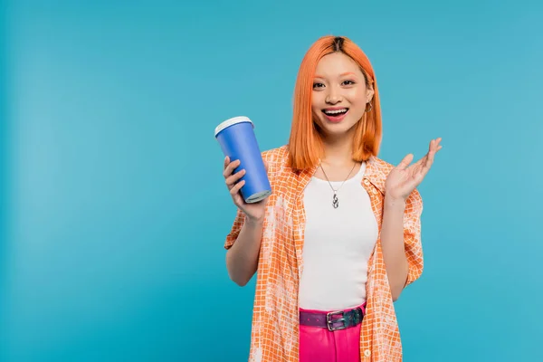 Positivity, drink in hand, happy asian and young woman with red hair holding paper cup and looking at camera on blue background, casual attire, generation z, coffee culture, hot beverage, amazed — Stock Photo