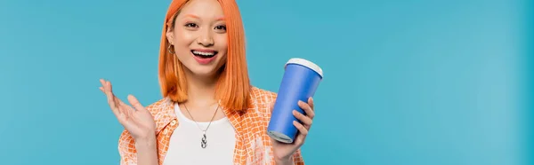 Positivité, café à emporter, heureuse asiatique et jeune femme aux cheveux roux tenant une tasse en papier et regardant la caméra sur fond bleu, tenue décontractée, génération z, culture du café, boisson chaude, étonné, bannière — Photo de stock
