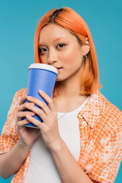 Modelo feminino beber café para ir, mulher asiática e jovem com cabelo vermelho segurando copo de papel e olhando para a câmera no fundo azul, traje casual, geração z, cultura do café, bebida quente — Fotografia de Stock