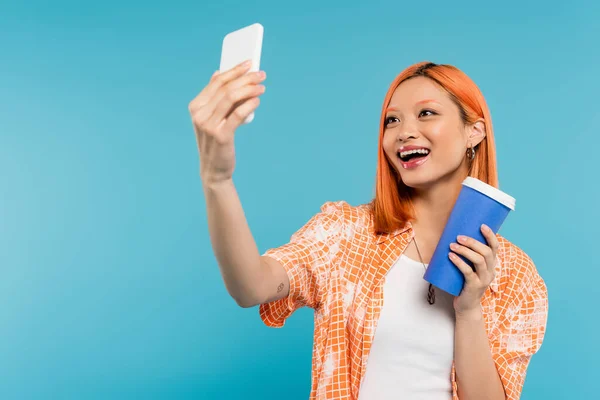 Tomando selfie, feliz asiático e jovem mulher com cabelo vermelho segurando copo de papel e usando smartphone em fundo azul, traje casual, geração z, cultura do café, influenciadores de mídia social — Fotografia de Stock
