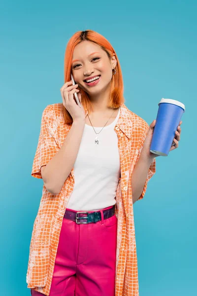 Телефонный звонок, веселая азиатская женщина с рыжими волосами держа кофе идти в бумажной чашке и говорить на смартфоне на голубом фоне, случайный наряд, поколение z, культура кофе, горячий напиток — стоковое фото