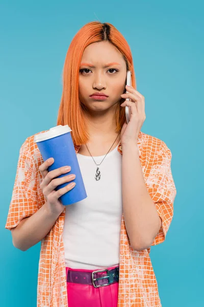 Phone call, upset asian woman with red hair holding coffee to go in paper cup and talking on smartphone on blue background, casual attire, generation z, coffee culture, displeased, pouting lips — Stock Photo