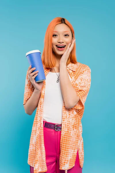 Positivität, erstaunte asiatische und junge Frau mit roten Haaren, die Kaffee to go in Pappbecher hält und in die Kamera auf blauem Hintergrund schaut, lässige Kleidung, Generation z, Kaffeekultur, Heißgetränk — Stockfoto