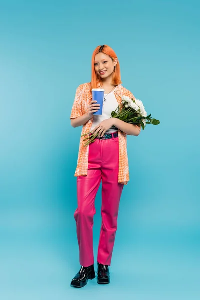 Full length of cheerful asian and young woman with red hair holding coffee to go and bouquet of flowers on blue background, casual attire, generation z, coffee culture, hot beverage, paper cup — Stock Photo