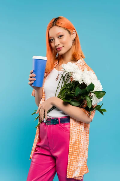 Весенние вибрации, азиатская и молодая женщина с красными рыжими волосами держа бумажную чашку и букет цветов на голубом фоне, случайный наряд, поколение z, культура выноса, горячий напиток, кофе на вынос — стоковое фото