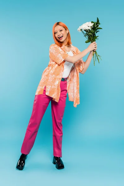 Positivité, femme asiatique étonnée aux cheveux roux tenant des fleurs blanches sur fond bleu, tenue décontractée, génération z, bouquet floral, vibrations printanières, visage heureux, gen z, culture de la jeunesse, pleine longueur — Photo de stock