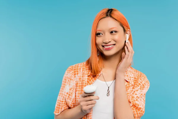 Bonheur d'été, amateur de musique, jeune femme asiatique à la mode avec des cheveux teints, dans un étui à chemise orange, réglage de l'écouteur sans fil et regardant loin sur fond bleu, génération z — Photo de stock