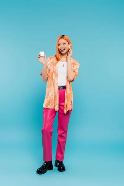 Full length of happy and stylish asian woman with colored red hair, in orange shirt and pink pants holding earphone case and listening music while standing on blue background in studio — Stock Photo