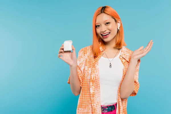Amazed asian woman with happy face and colored red hair wearing stylish orange shirt and listening music in wireless earphone while standing with case on blue background, youth culture — Stock Photo