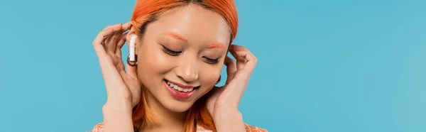 Happy face, radiant smile, young asian woman fixing dyed red hair while listening music in wireless earphone on blue background, summer vibes, youth culture, banner — Stock Photo