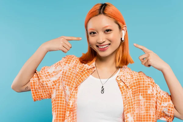 Été insouciant, femme asiatique à la mode avec les cheveux roux et en chemisier orange pointant avec les doigts à l'écouteur, écouter de la musique et en regardant la caméra sur fond bleu, tatouage, génération z style de vie — Photo de stock