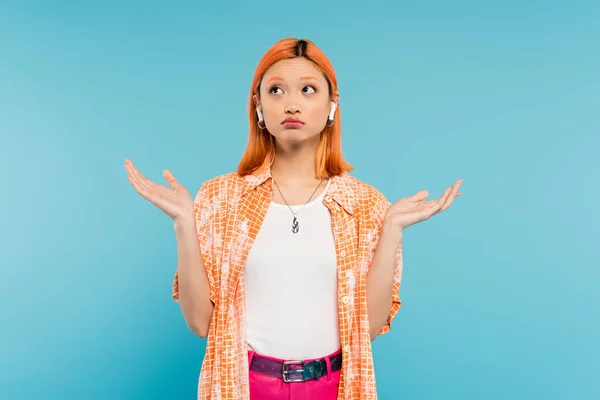 Jeune femme asiatique découragée en chemise orange, avec des cheveux roux teints écoutant de la musique dans un écouteur sans fil, montrant un geste haussant les épaules et regardant vers le haut sur fond bleu, génération z — Photo de stock