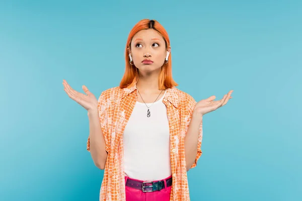 Giovane e scoraggiata donna asiatica in camicia arancione, con capelli rossi tinti ascoltare musica in auricolare wireless, mostrando gesto scrollata di spalle e guardando su sfondo blu, generazione z — Foto stock