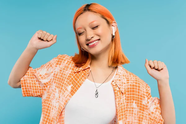 Glück, Musikliebhaberin, junge Asiatin mit gefärbten roten Haaren, im trendigen orangefarbenen Hemd und drahtlosem Kopfhörer gestikulierend mit geschlossenen Augen auf blauem Hintergrund, Generation-Z-Lifestyle, Sommerstimmung — Stockfoto