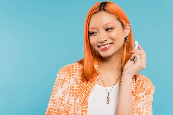 Young and happy asian fashion model with colored red hair, in stylish orange shirt holding wireless earphone while standing on blue background, youth culture, generation z — Stock Photo
