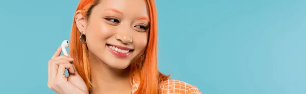 Bonheur et loisirs, portrait d'une adorable femme asiatique avec un sourire radieux et des cheveux roux colorés tenant un écouteur sans fil sur fond bleu, vibrations estivales, génération z, bannière — Photo de stock