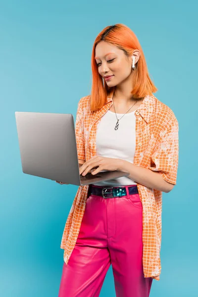 Émotion positive, jeune femme asiatique à la mode avec un visage souriant debout sur fond bleu et travaillant sur ordinateur portable, chemise orange, pantalon rose, mode moderne, mode de vie freelance — Photo de stock