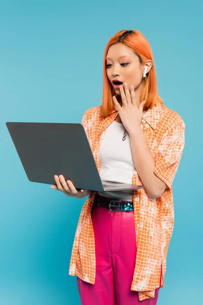Staunende asiatische Frau mit rot gefärbten roten Haaren, die die Hand in der Nähe des offenen Mundes hält und auf den Laptop auf blauem Hintergrund schaut, jugendliche Mode, orangefarbenes Hemd, freiberuflicher Lebensstil — Stockfoto