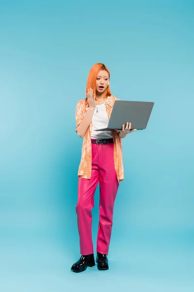 Überraschte Emotion, Videoanruf, rothaarige asiatische Frau mit offenem Mund in orangefarbenem Hemd und pinkfarbener Hose, die neben Laptop auf blauem Hintergrund wedelt, freiberuflicher Lebensstil, Generation z — Stockfoto