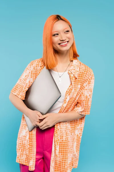 Felicidad de verano, atractiva mujer asiática con el pelo rojo y sonrisa radiante sosteniendo el ordenador portátil y mirando hacia otro lado mientras está de pie en camisa naranja sobre fondo azul, estilo de vida independiente, generación z - foto de stock