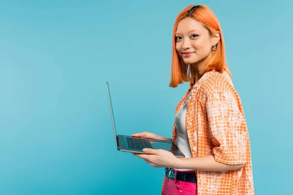 Glückliches Gesicht, optimistische Emotion, jugendliche Asiatin mit gefärbten roten Haaren, im orangefarbenen Hemd Laptop in der Hand und Kamera auf blauem Hintergrund, freiberuflicher Lebensstil, Generation z — Stockfoto
