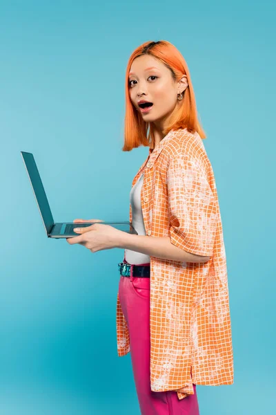 Étonnement, surprise, jeune femme asiatique avec des cheveux rouges de couleur rouge et bouche ouverte tenant ordinateur portable et regardant la caméra sur fond bleu, mode jeune, chemise orange, mode de vie freelance — Photo de stock