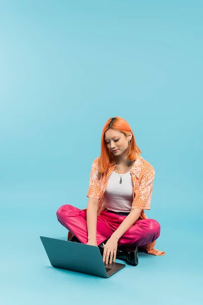 Femme asiatique à la mode en pantalon rose et chemise orange travaillant sur ordinateur portable tout en étant assis avec les jambes croisées sur fond bleu, souriant, émotion positive, mode de vie freelance, génération z — Photo de stock