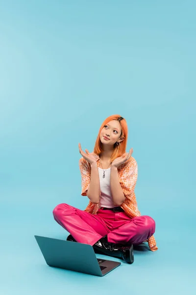 Émotion positive, femme asiatique rêveuse et souriante assise avec les jambes croisées près d'un ordinateur portable sur fond bleu, cheveux rouges colorés, chemise orange, pantalon rose, mode de vie freelance, génération z — Photo de stock